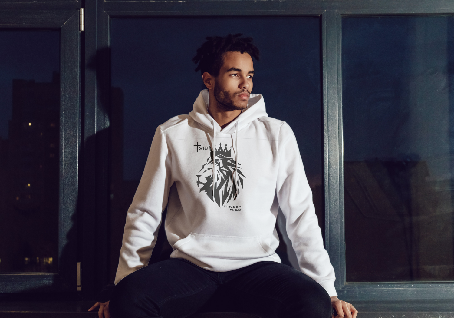 An African American man wearing a hoodie sits on a window ledge, gazing thoughtfully outside.