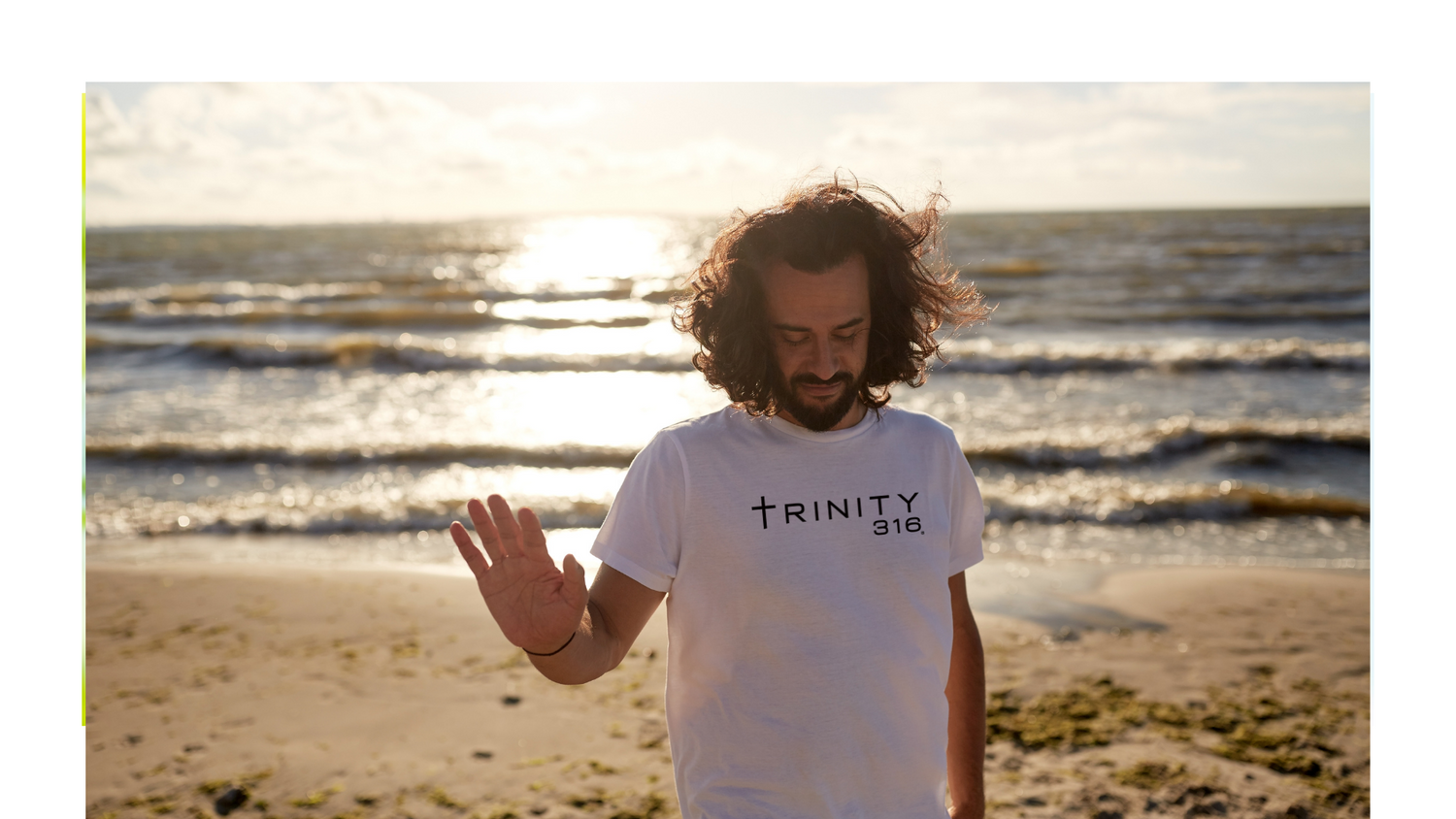 A man with long hair wearing a white t-shirt waves cheerfully at the camera, showcasing a friendly demeanor.