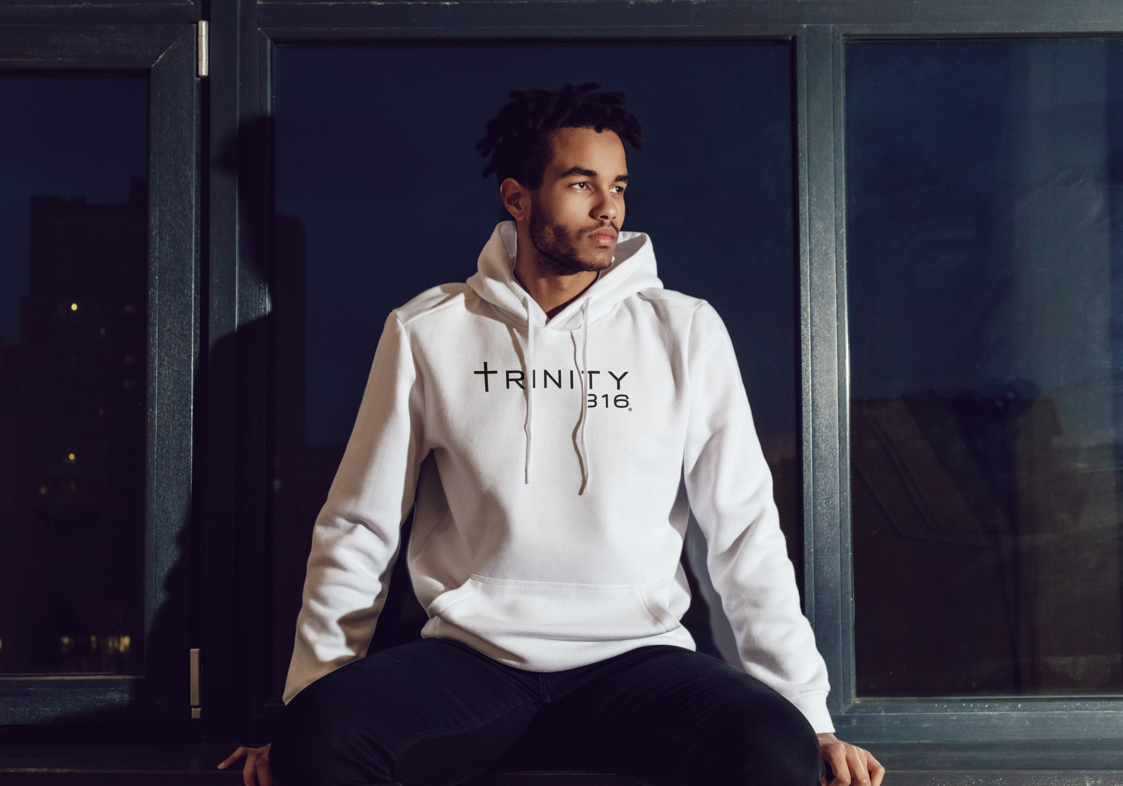An African American man in a white hoodie sits on a window ledge, with the Trinity 316 logo visible nearby.