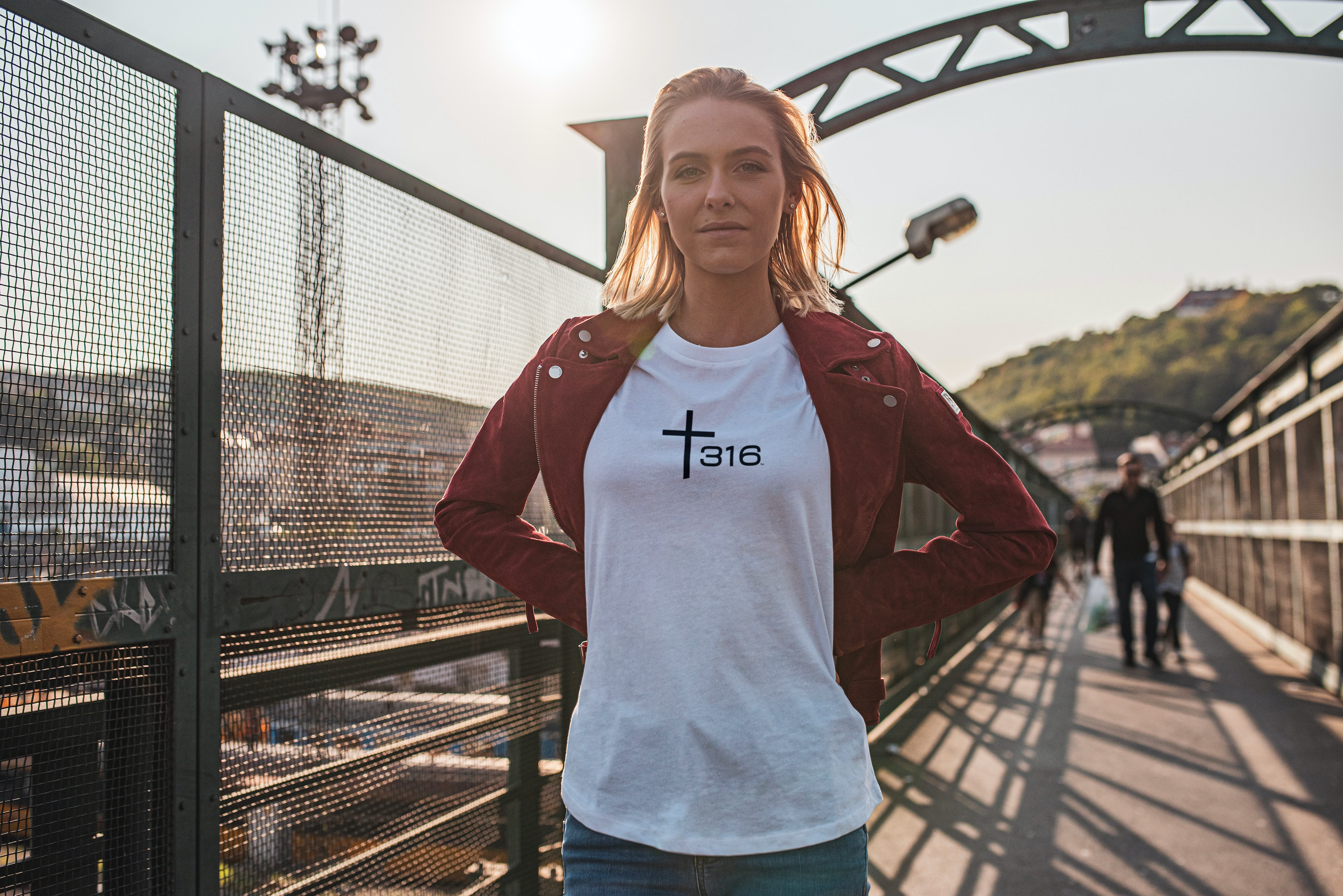 A blonde woman stands on a bridge, wearing a T-shirt that reads faith, with the Trinity316 icon in black.